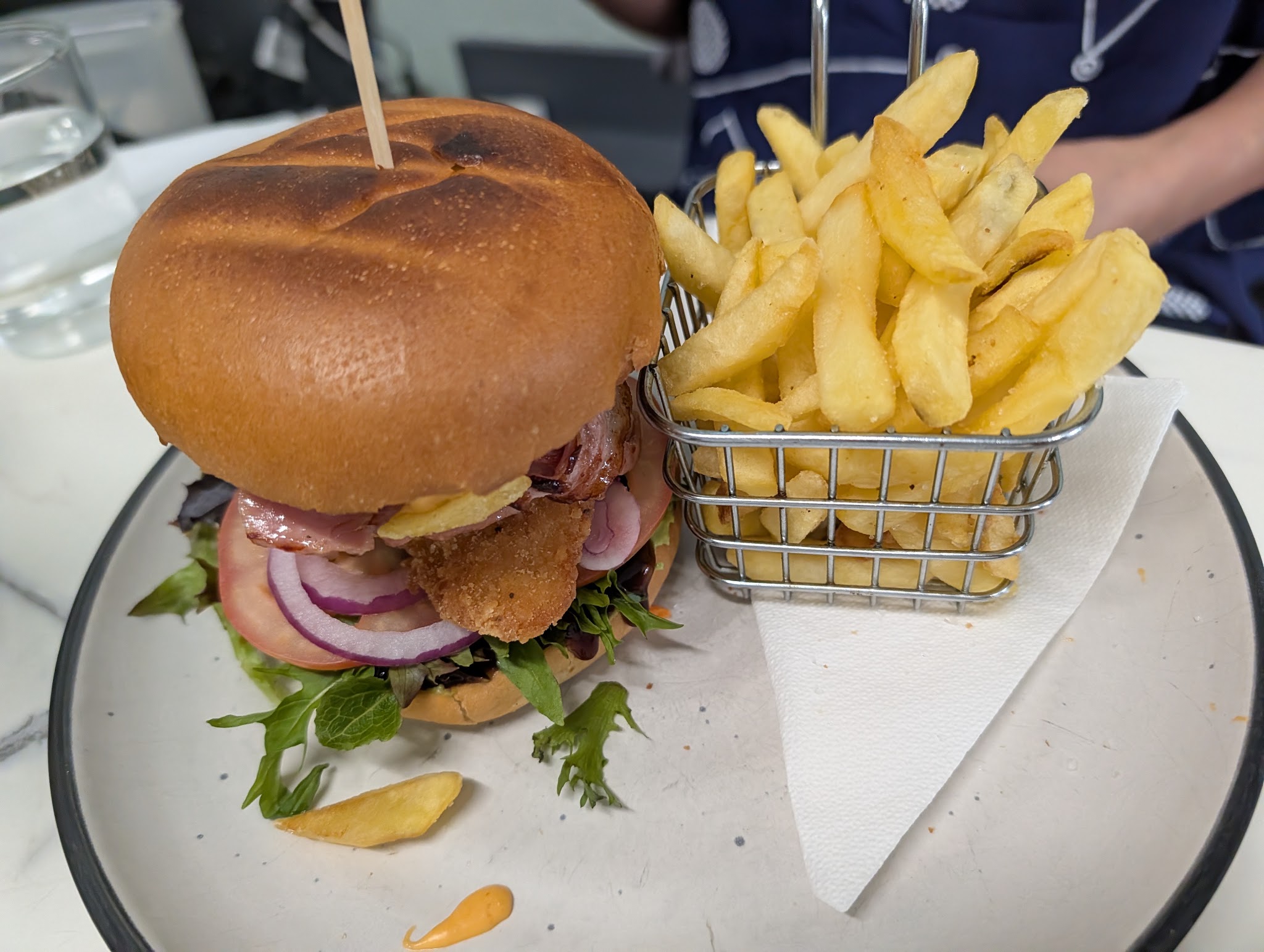 Burger with bacon, crumbed chicken and salad beside a basket of chips